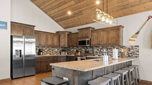 A modern kitchen with wooden cabinets, stainless steel appliances, a large island with bar stools, and a unique ceiling design. - 2BR Retreat Near SDC w/ Hot Tub + Boat Slip (Branson)