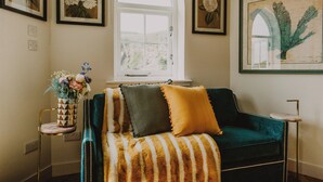 Een gezellige kamer met een groene fluwelen bank, een gestreept plaid en een klein bijzettafeltje met een vaas vol bloemen.