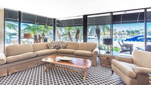 A modern living room with a large window, a sofa, a coffee table, and a view of a marina. - Charming 5BR Mid-Century Bayfront Retreat with Floor-to-Ceiling Windows! (Newport Beach)