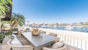A patio with wicker furniture, a table, and a view of a marina with boats. - Charming 5BR Mid-Century Bayfront Retreat with Floor-to-Ceiling Windows! (Newport Beach)