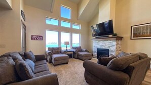A spacious living room with a stone fireplace, a large flat-screen TV, and a view of the outdoors through large windows.