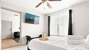 Een slaapkamer met een groot bed, een plafondventilator, een schilderij aan de muur en een raam met gordijnen.