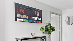 Een woonkamer met een tv aan de muur waarop streamingdiensten te zien zijn, een console tafel met een klok, een potplant en een ronde glazen tafel met een zwart-wit vloerkleed.