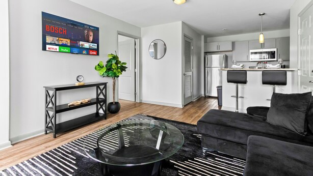 Een moderne woonkamer met een glazen salontafel, een zwarte bank en een gestreept vloerkleed. Een flatscreen televisie aan de wand toont diverse streamingdiensten.