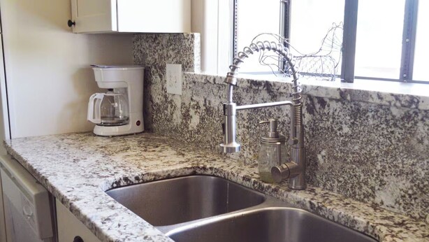 Cucina con piani di lavoro in granito, lavelli in acciaio inossidabile, macchina da caffè e finestra con zanzariera.