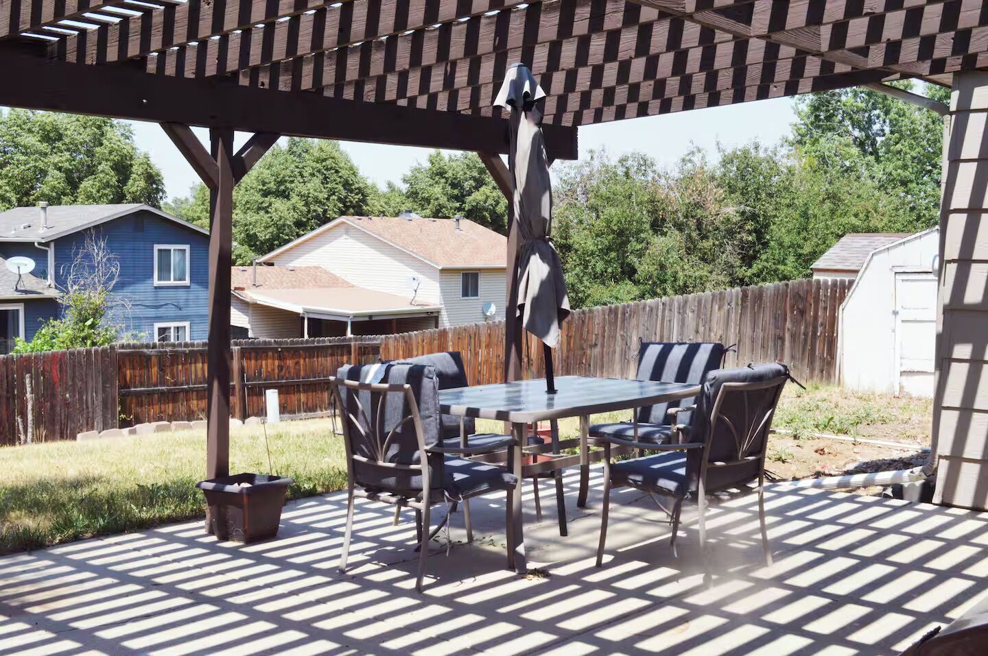 Een terras buiten met een pergola, een tafel met stoelen en een hangende lamp.