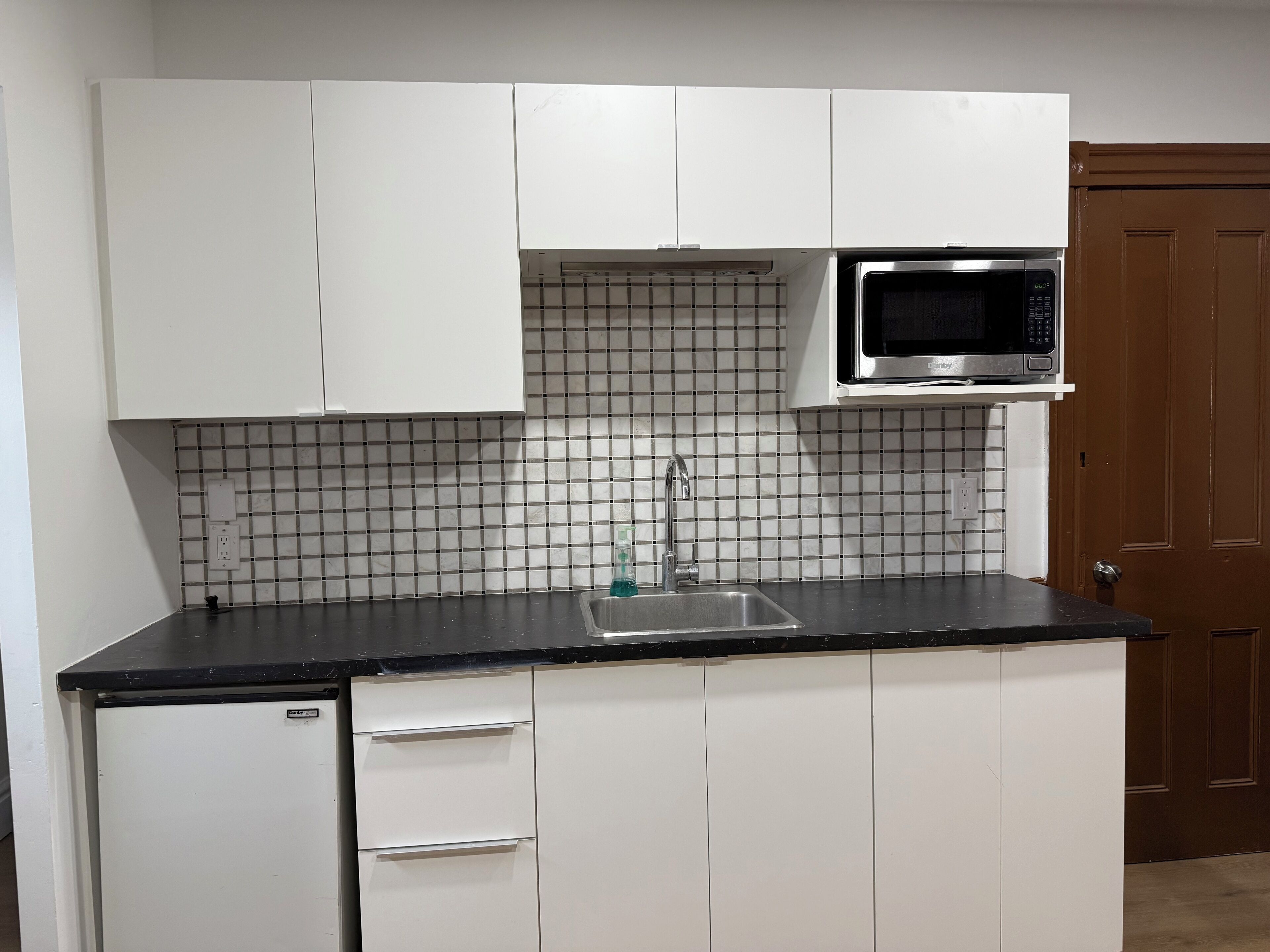A compact kitchen with white cabinets, a stainless steel sink, a microwave, and a tiled backsplash.
