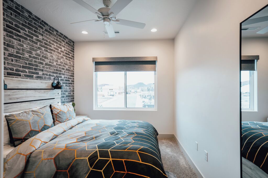 A bedroom with a brick wall, a large window, a ceiling fan, and a bed with patterned bedding.