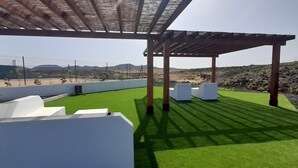 Un espacio exterior techado con césped artificial verde, macetas blancas y vistas a montañas distantes.