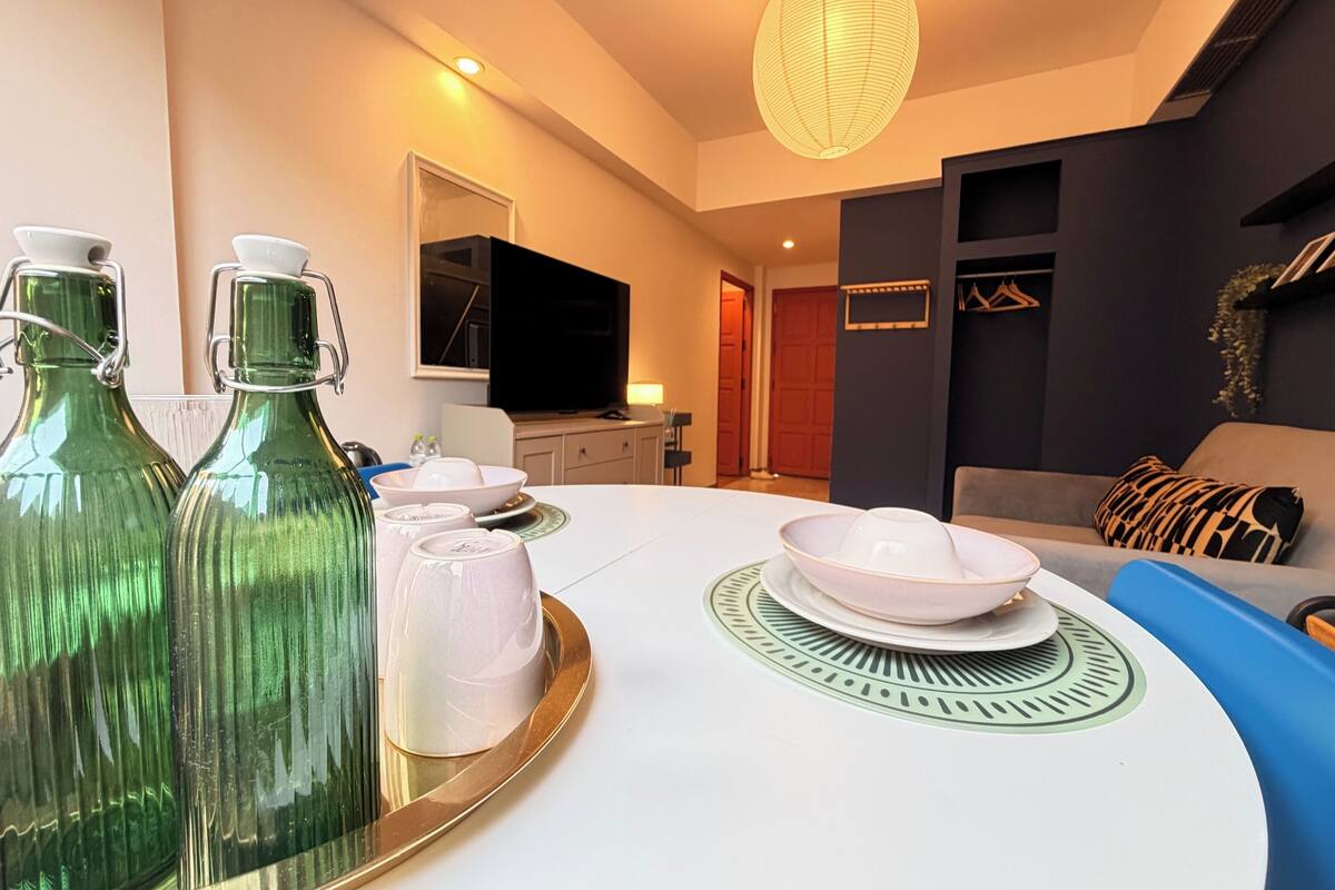A modern living room with a round dining table, green glass bottles, white plates, and a flat-screen TV.