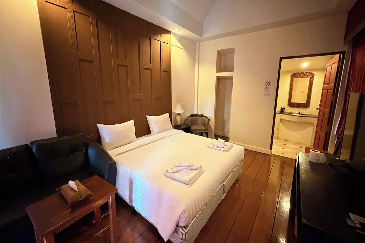 A hotel room with a bed, a wooden headboard, a small table with a tissue box, a chair, and a mirror in the bathroom.