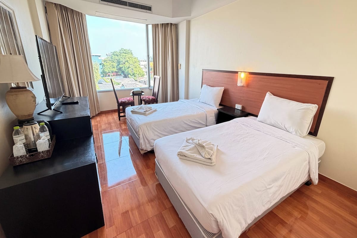 A hotel room with two beds, a desk with a lamp, a chair, and a window with curtains.