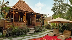 A traditional wooden building with a tiled roof, surrounded by a garden with a red flower bed and two lounge chairs under an umbrella. - Hidden Saren Villa Ubud (Ubud)