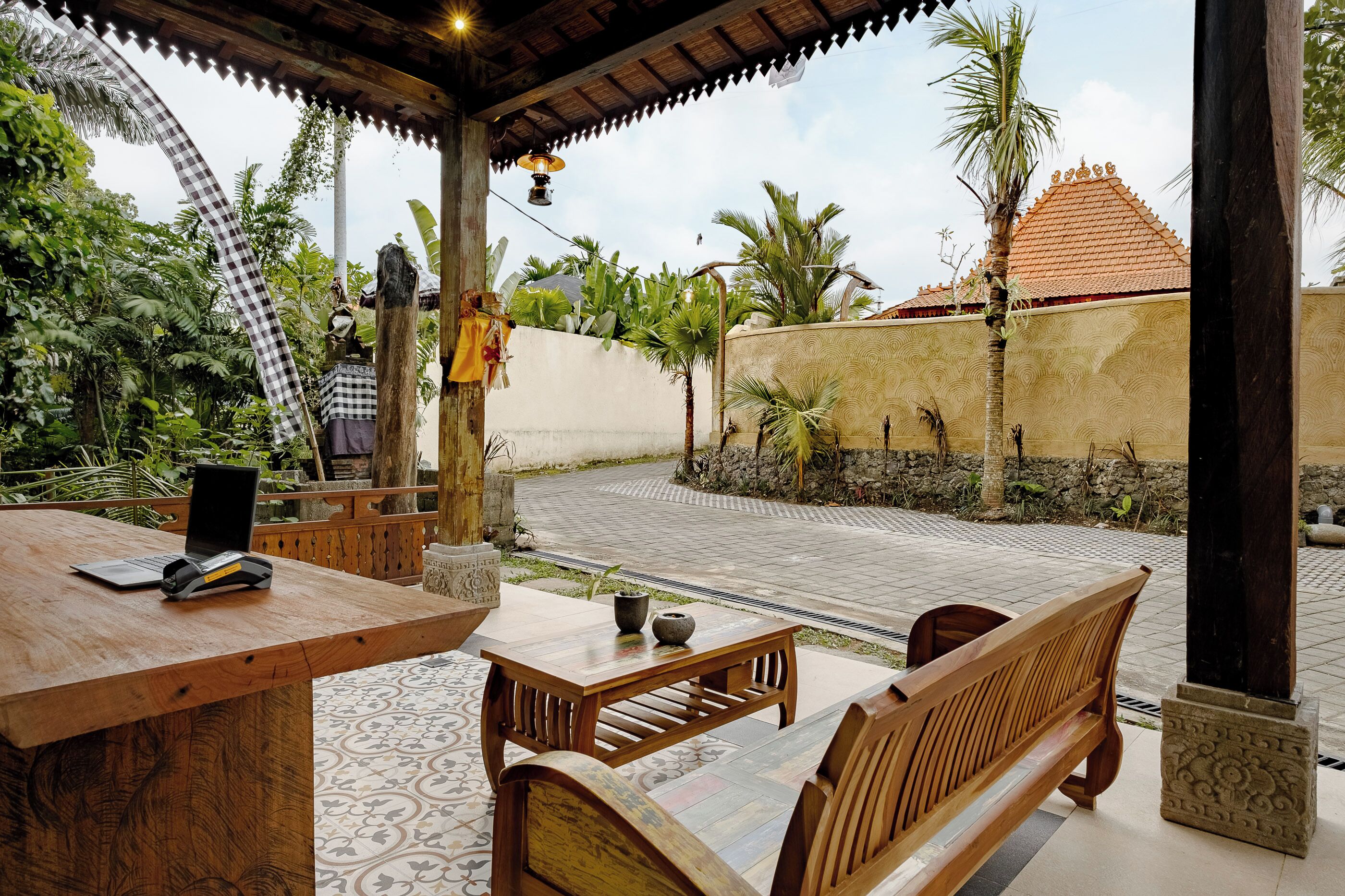 An outdoor seating area with wooden furniture, a table with a laptop and a vase, surrounded by tropical plants and a decorative wall.