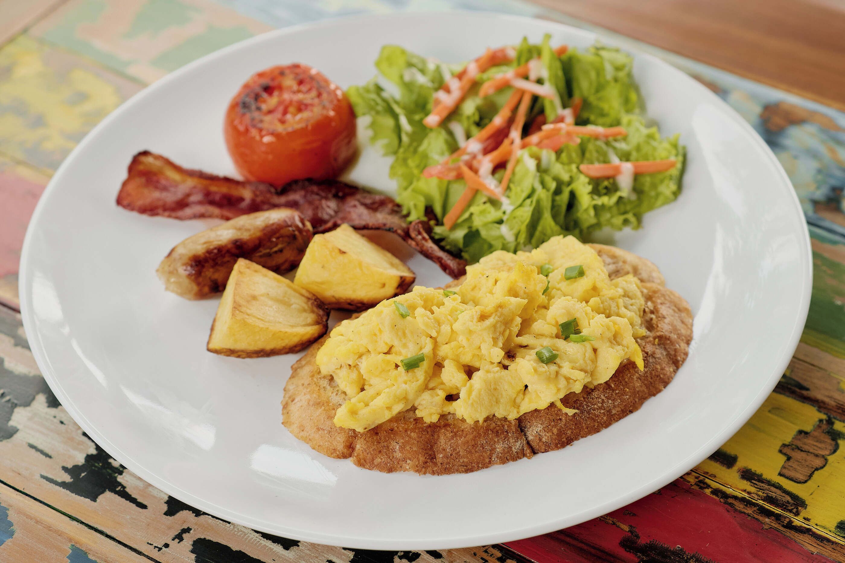 A plate with scrambled eggs, bacon, roasted potatoes, a grilled tomato, and a side salad.