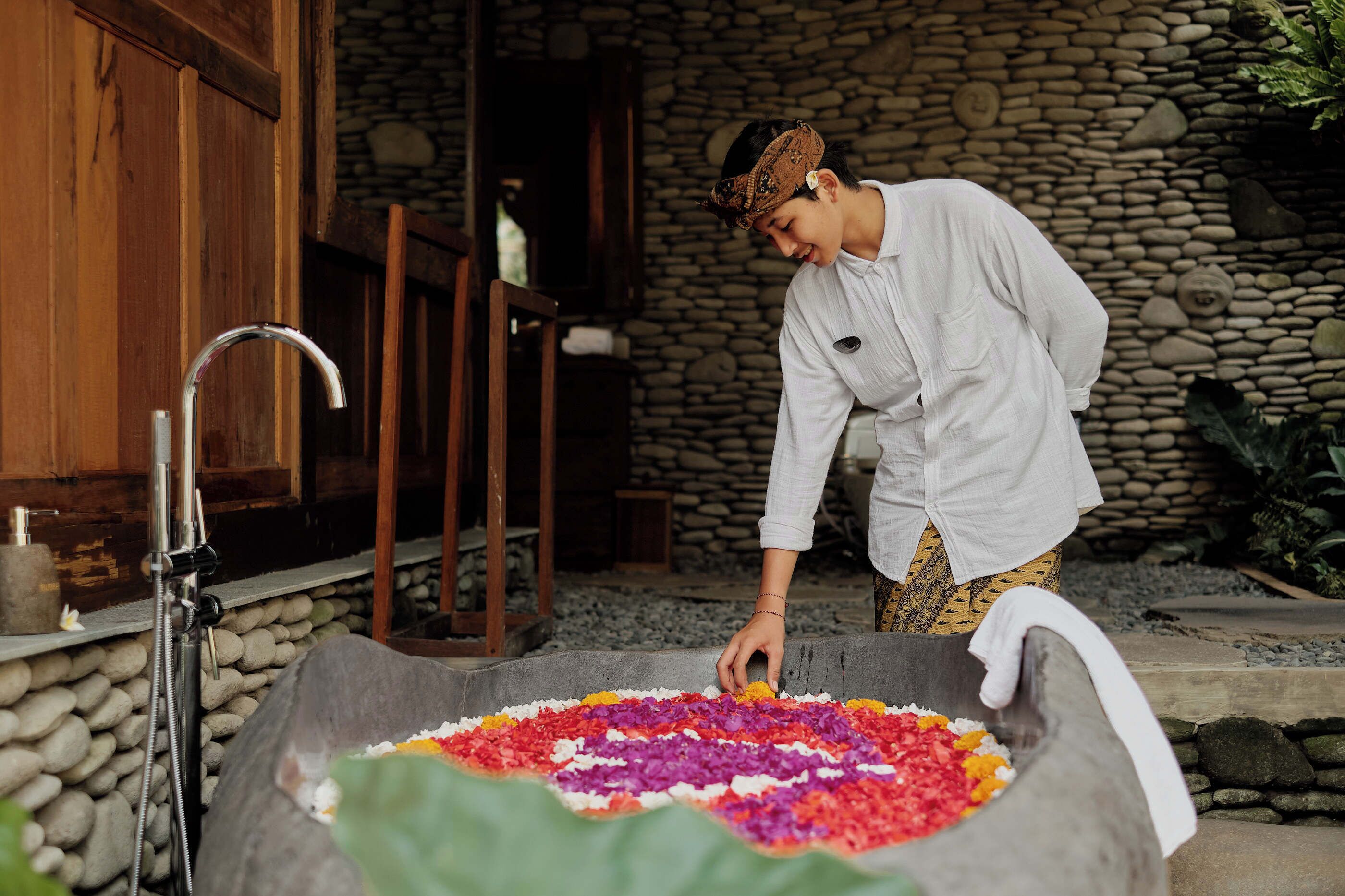 A person arranging colorful flowers in a stone basin outdoors, with a stone wall and wooden furniture in the background.