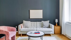 A living room with a grey sofa, two pillows, a round coffee table with books, a pink chair, and a small wooden side table with a lamp.