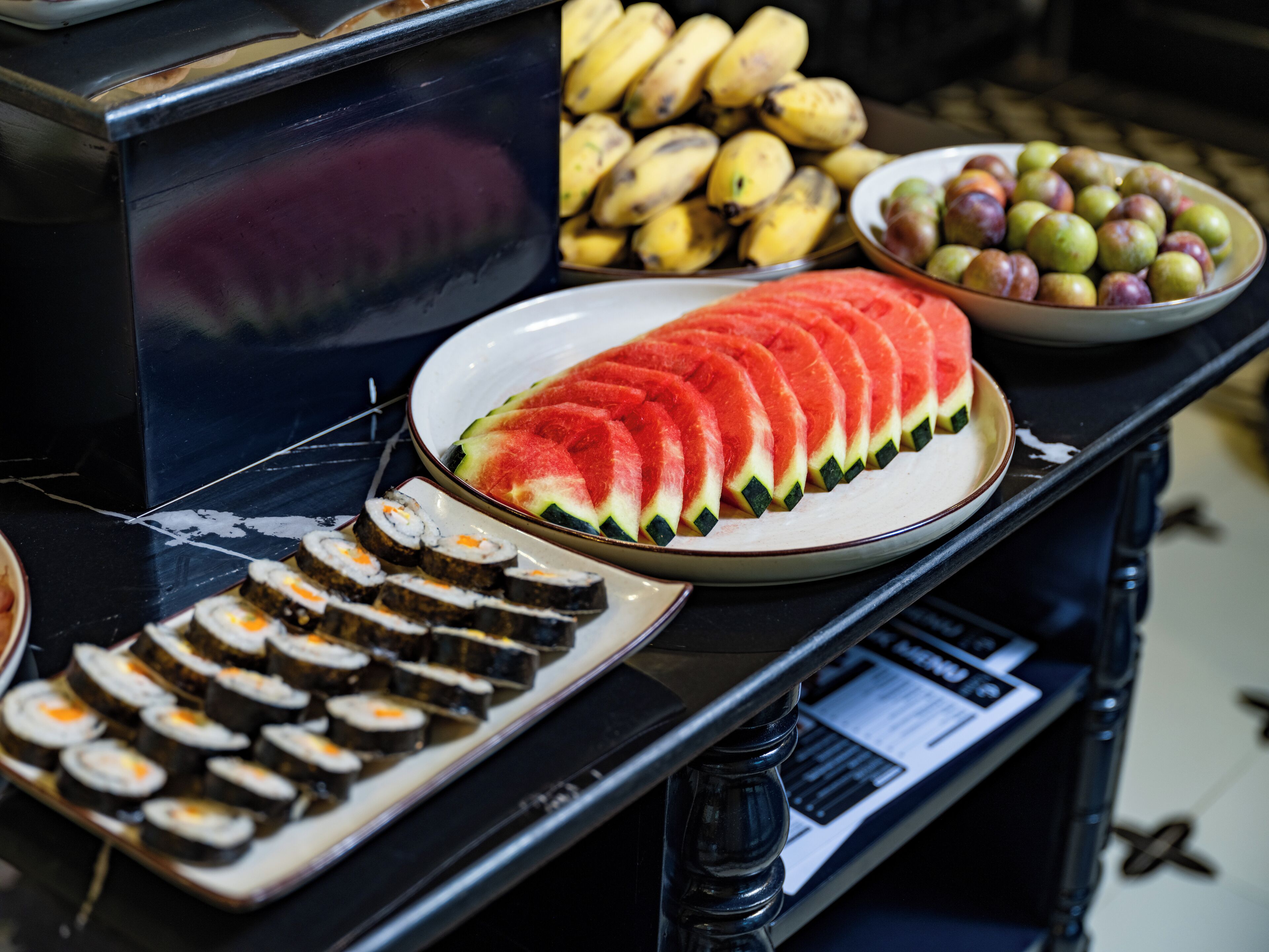 A buffet table with sliced watermelon, bananas, and a plate of sushi.