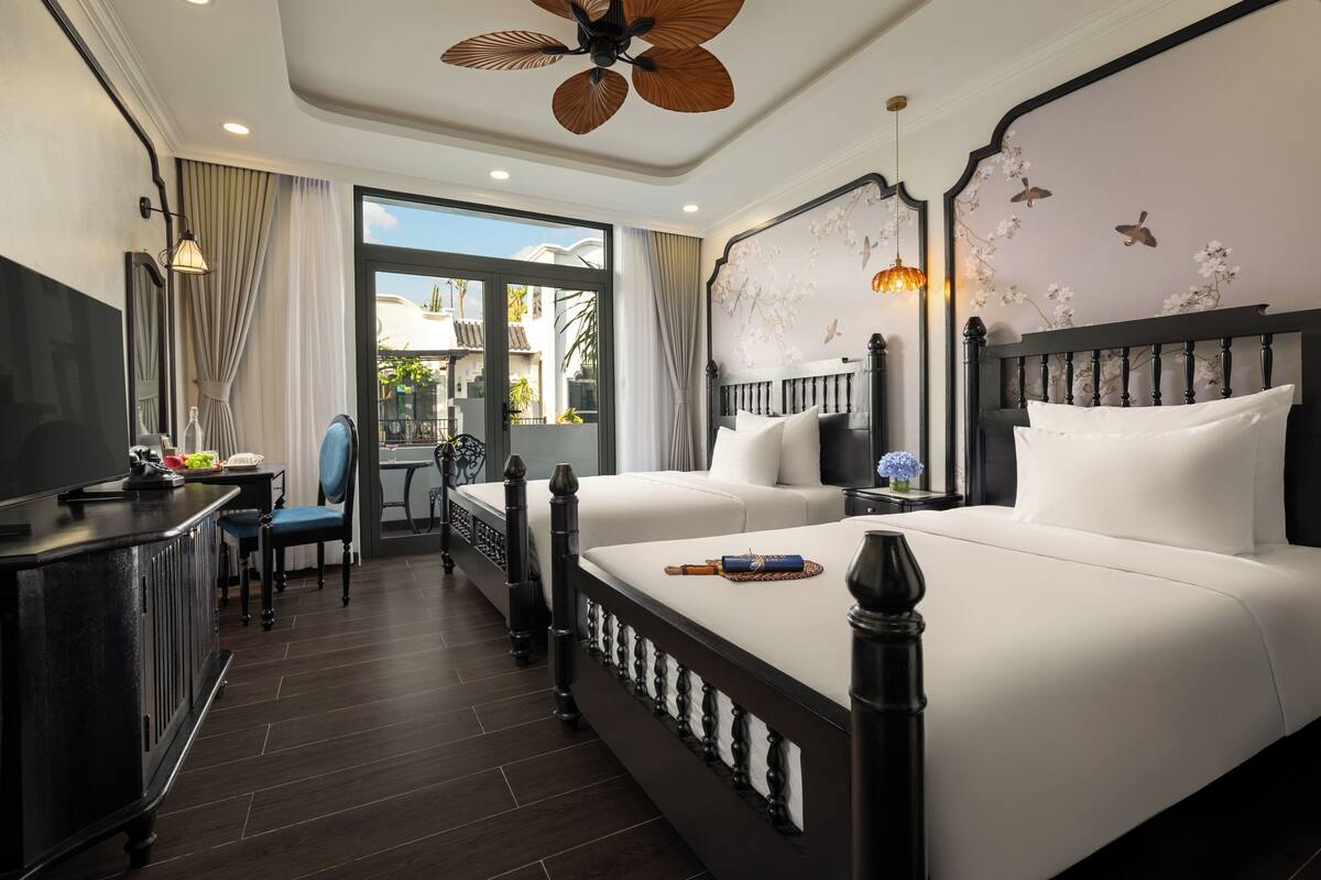 A hotel room with a large bed, a desk with a chair, a television, and a balcony with curtains.