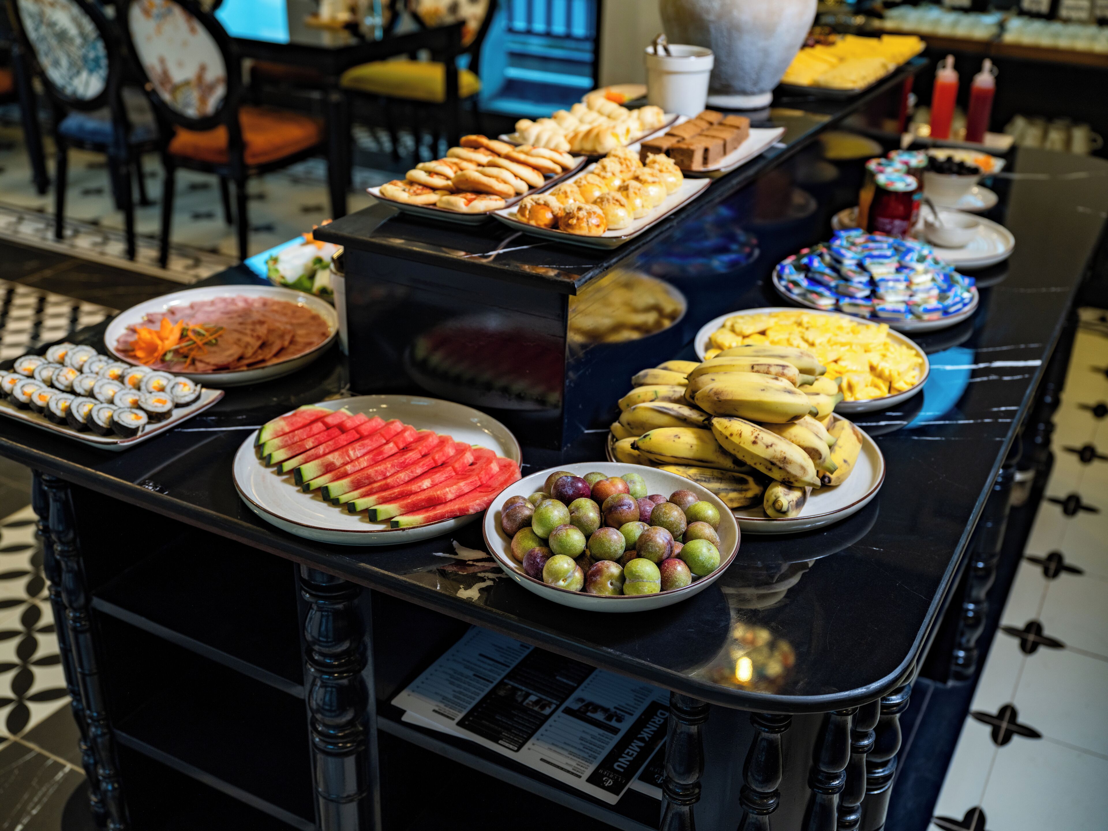 A buffet table with various food items including sushi, watermelon slices, bananas, and pastries.