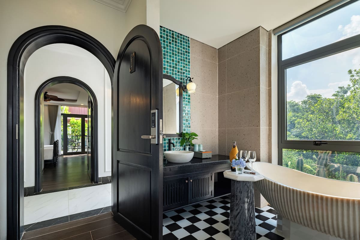 A modern bathroom with a black and white checkered floor, a large window with a view of greenery, and a black door with a light switch.