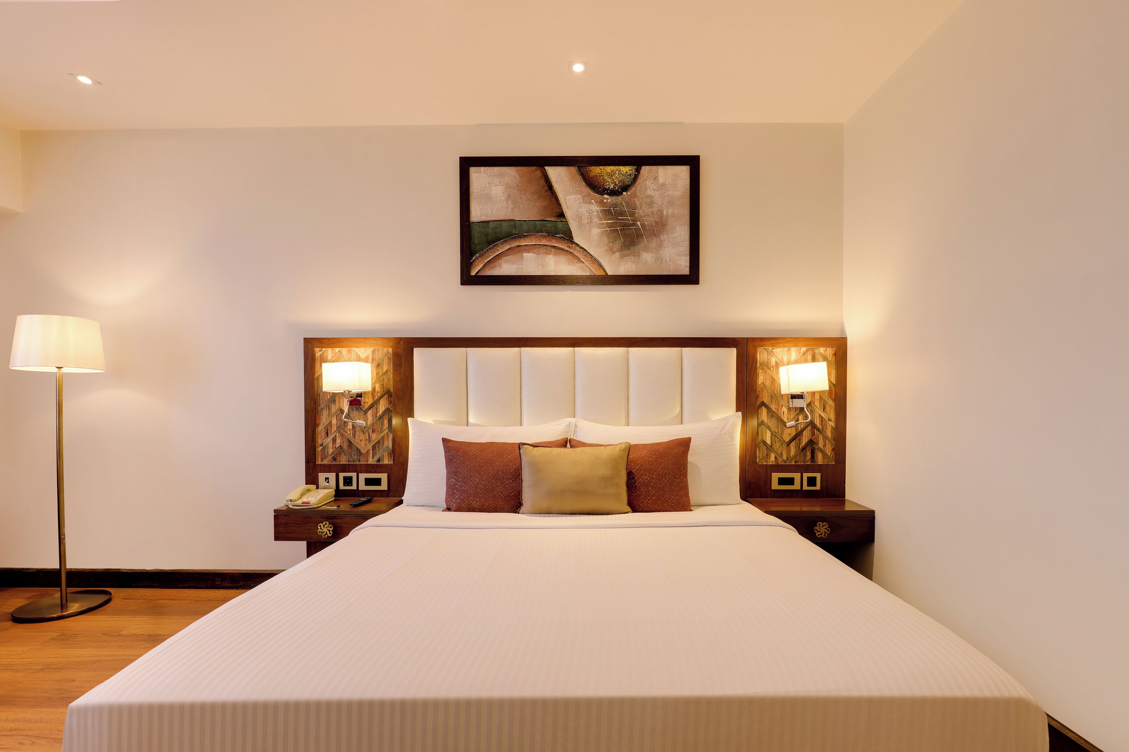 Una habitación de hotel con una cama grande, cabecera con panel de luz, lámparas de noche, un cuadro colgado en la pared y una lámpara de pie.