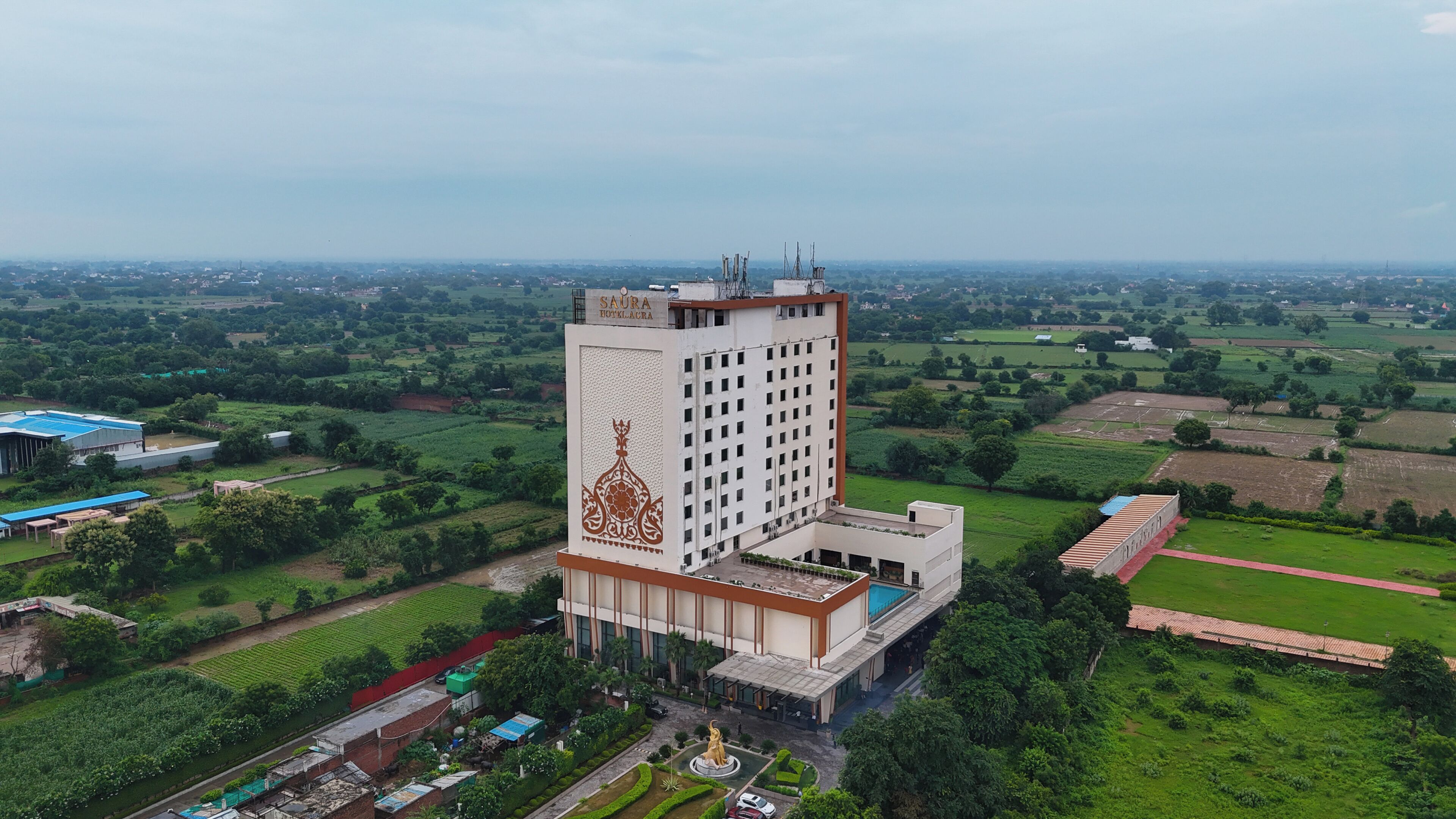 A high-rise hotel with a central courtyard, surrounded by green fields and a distant cityscape.
