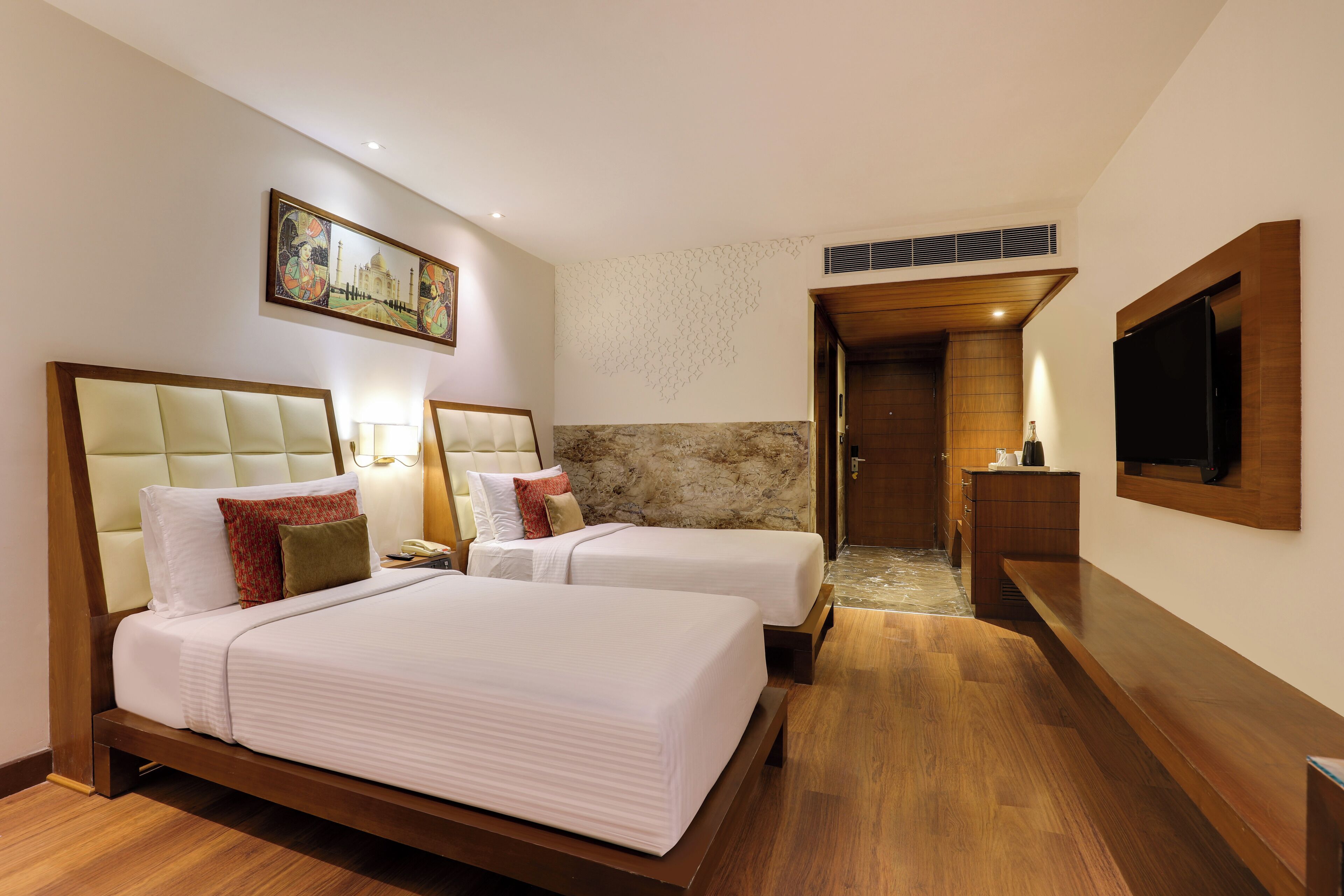 A hotel room with two beds, a wooden floor, a television, and a painting on the wall.