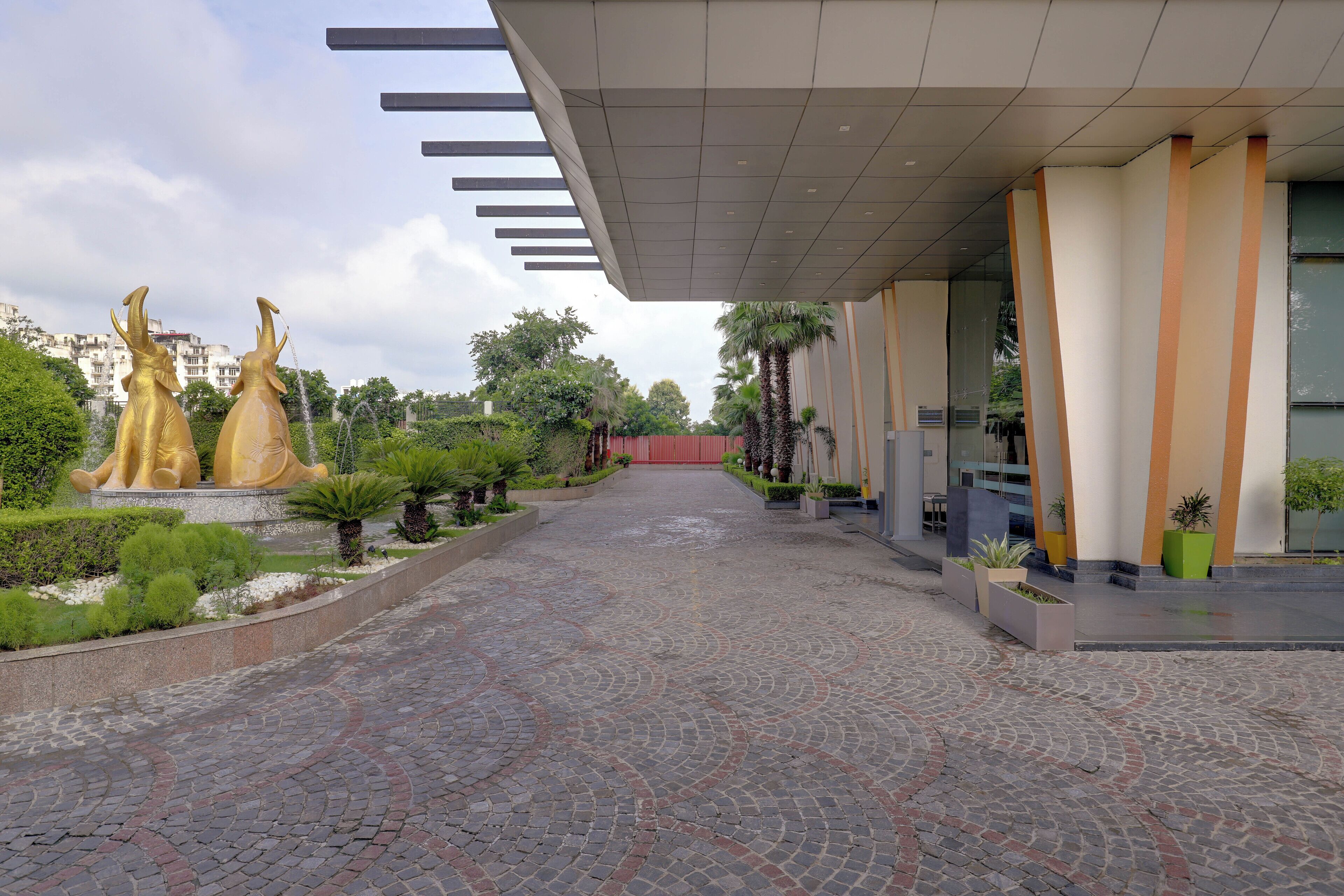 Un percorso lastricato conduce a un edificio dal design architettonico moderno, caratterizzato da una serie di statue dorate e aree verdi curate.