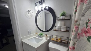A bathroom with a white sink, a round mirror, a toilet, and a shower curtain with floral patterns. - Charming studio ,minutes from the airport. (Hialeah)