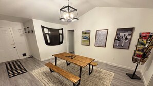 A room with a wooden table and bench, a patterned rug, a wall-mounted light fixture, and framed pictures.