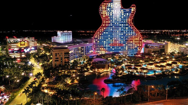 Un hotel con fachada en forma de guitarra, iluminado con luces rojas y azules, rodeado de una zona de piscina y palmeras.