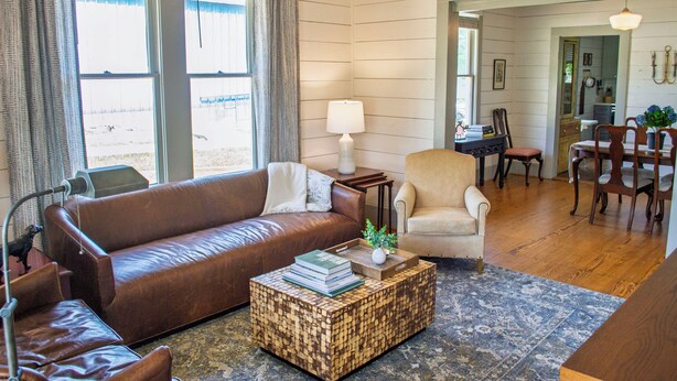 A cozy living room with a brown leather sofa, a small wooden coffee table with books, a white lamp, and a dining area with wooden chairs and a table. - Immaculately restored home in Downtown Brenham (Brenham)