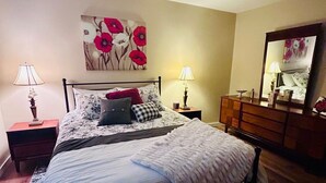 Un dormitorio con cama, dos mesitas de noche, cómoda, espejo y un cuadro floral en la pared.