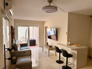 Uma sala de estar moderna com um sofá, uma mesa de centro e uma televisão na parede. Há uma porta de vidro deslizante que dá para uma área externa com um pátio.