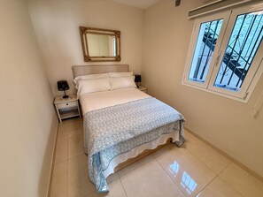 Quarto com cama, mesa de cabeceira, luminária, espelho e janela com vista para uma varanda.