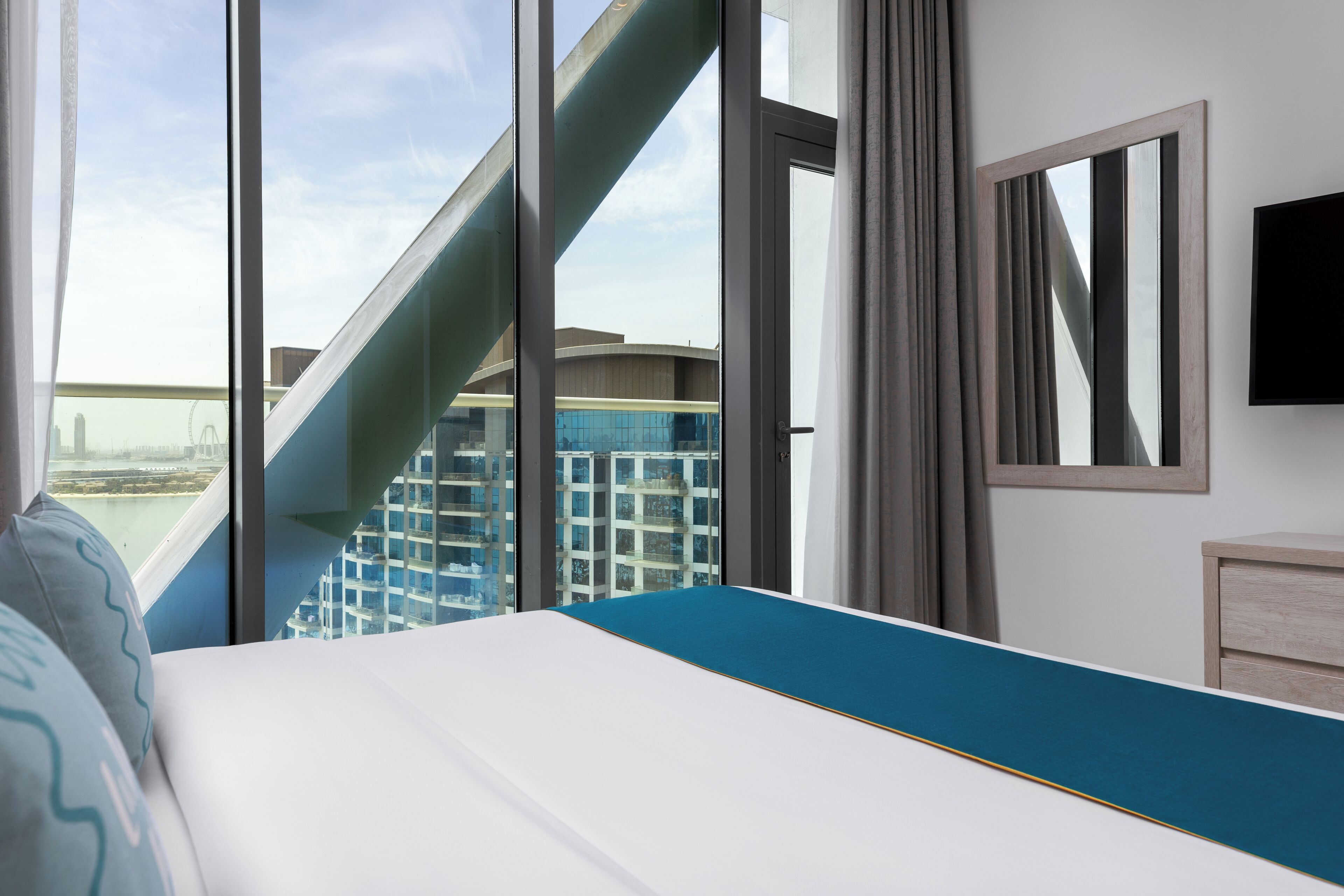Une chambre d’hôtel moderne dotée d’un grand lit, offrant une vue sur le paysage urbain et disposant d’un balcon avec une balustrade en verre.