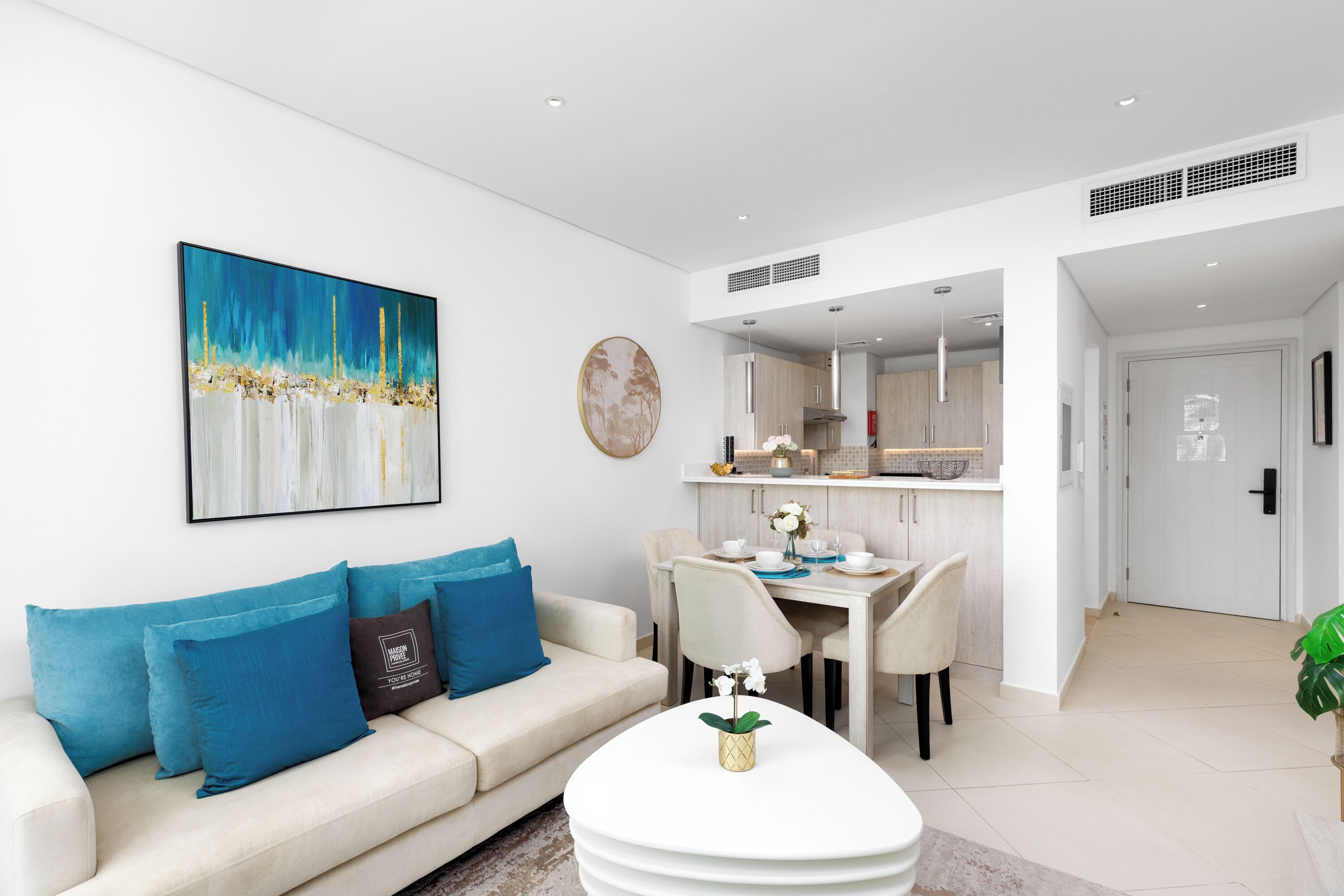 A modern living room with a sofa, coffee table, and artwork. A dining area with a table and chairs is visible in the background.