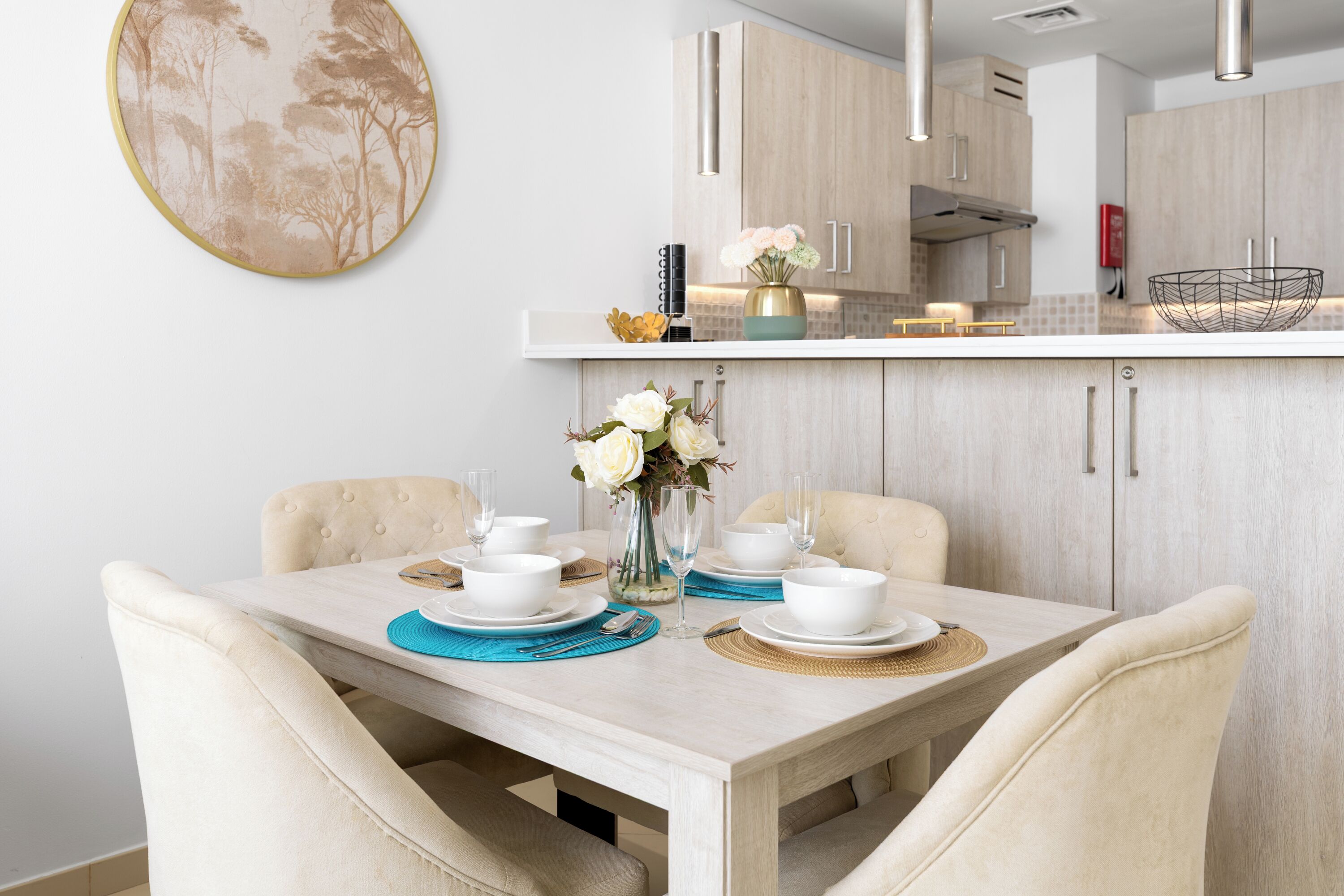 A dining area with a table set for two, a vase with flowers, and a kitchen in the background.
