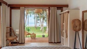 Ein Schlafzimmer mit einem großen Bett, einem Schaukelstuhl aus Holz, einem Korbstuhl und Blick auf den Strand und Palmen.