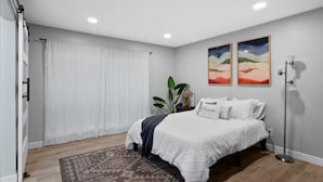 A bedroom with a bed, a large window with curtains, a floor lamp, and two wall-mounted paintings.