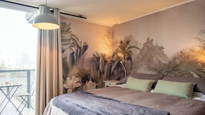 Une chambre dotée d’un grand lit, d’une table de chevet, d’une lampe, d’un balcon avec une table et des chaises, et d’une fresque représentant une montagne et du feuillage sur le mur.