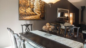 Een eetruimte met een donker houten tafel, stoelen, een groot schilderij met bosmotieven, een kroonluchter en een spiegel aan de muur.