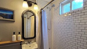 A bathroom with a white sink, a mirror, and a shower with a glass door. There is a window above the shower and a picture on the wall.