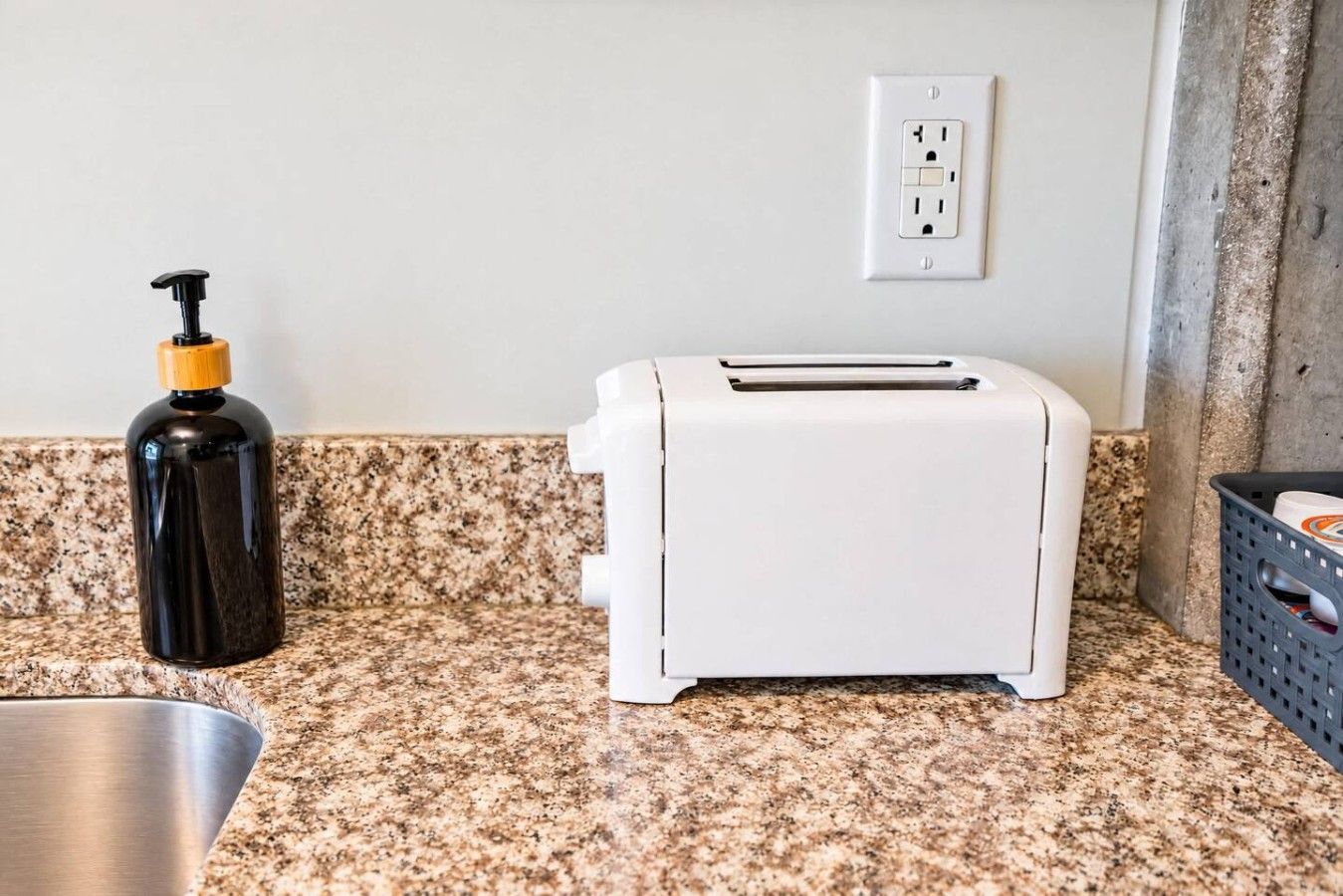A kitchen with a granite countertop, a toaster, a black bottle with a pump dispenser, and a power outlet on the wall.