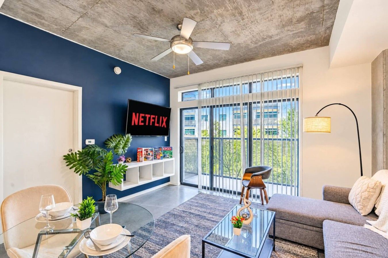 A modern living room with a flat-screen TV, a ceiling fan, and a large window with a view of buildings.