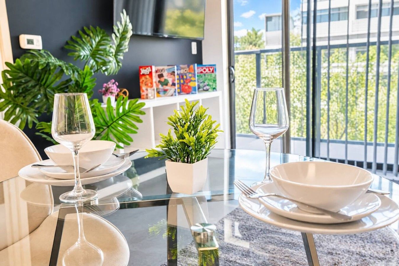 A dining area with a glass table, white plates, and wine glasses. There are potted plants and a bookshelf with books and games.