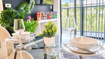 A dining area with a glass table, white plates, and wine glasses. There are potted plants and a bookshelf with books and games.