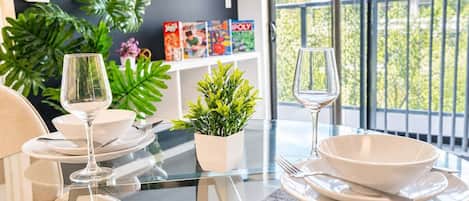 Un comedor con una mesa de vidrio, platos blancos y copas de vino. Hay plantas en macetas y una estantería con libros y juegos.