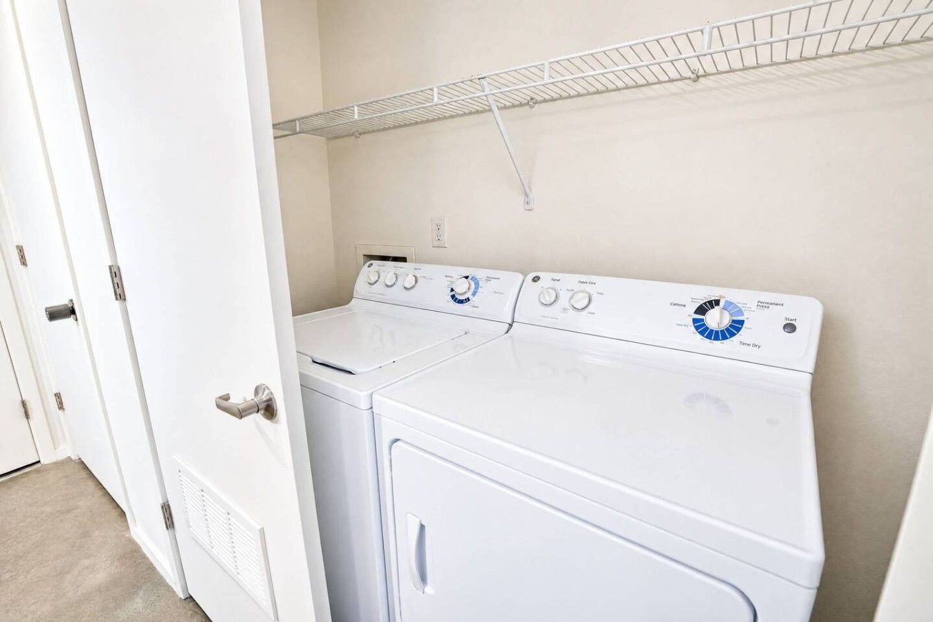 A laundry room with a front-loading washer and dryer, a metal shelf above, and a white door.